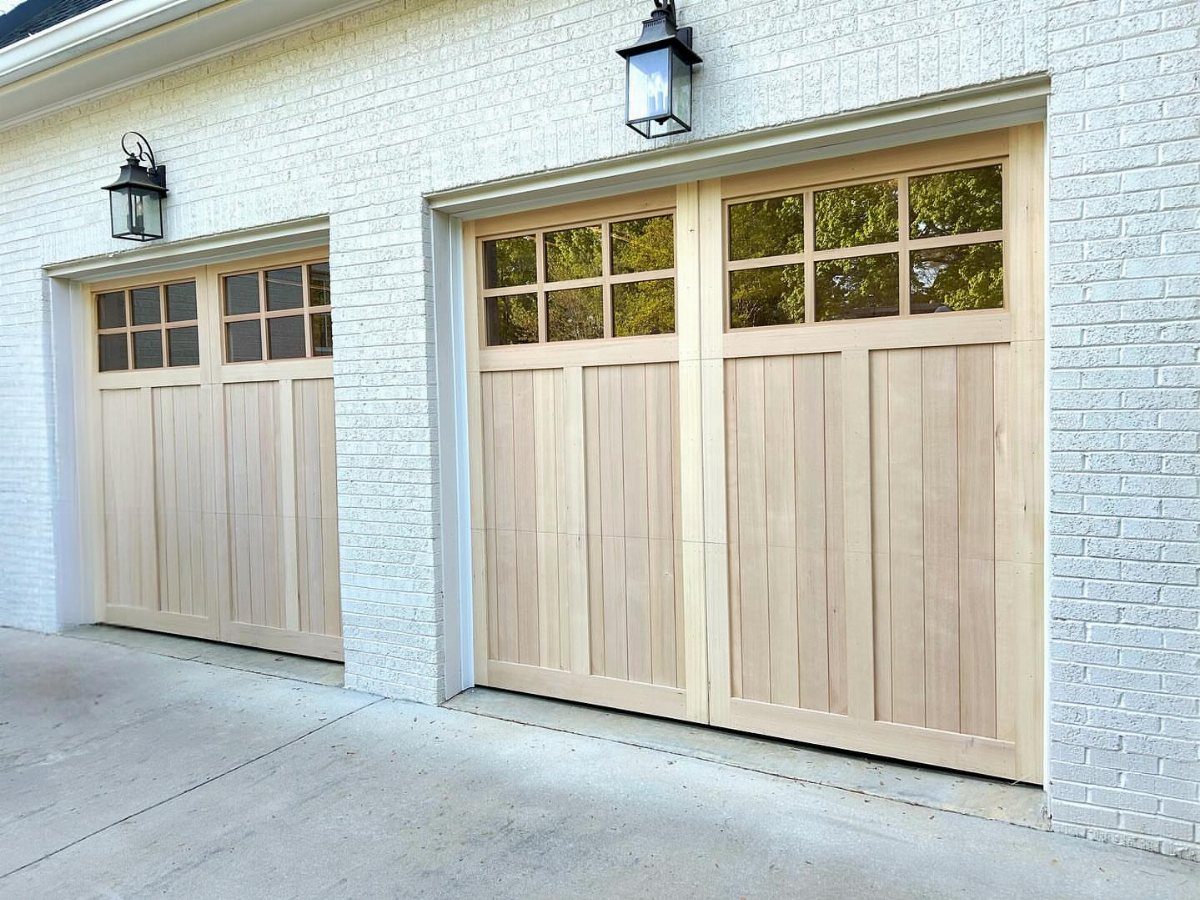 American House Garage Door: 7 Styles for Better Curb Appeal 3 Two light wood garage doors with window panes installed on a white brick house.