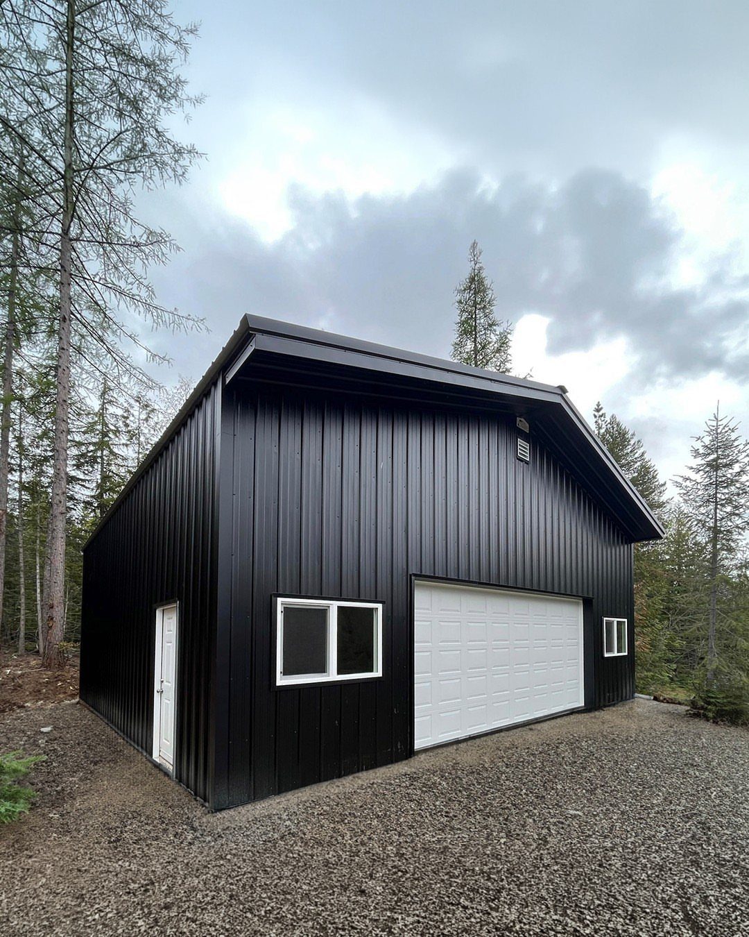 A black metal pole barn garage with white doors and windows in a forest.