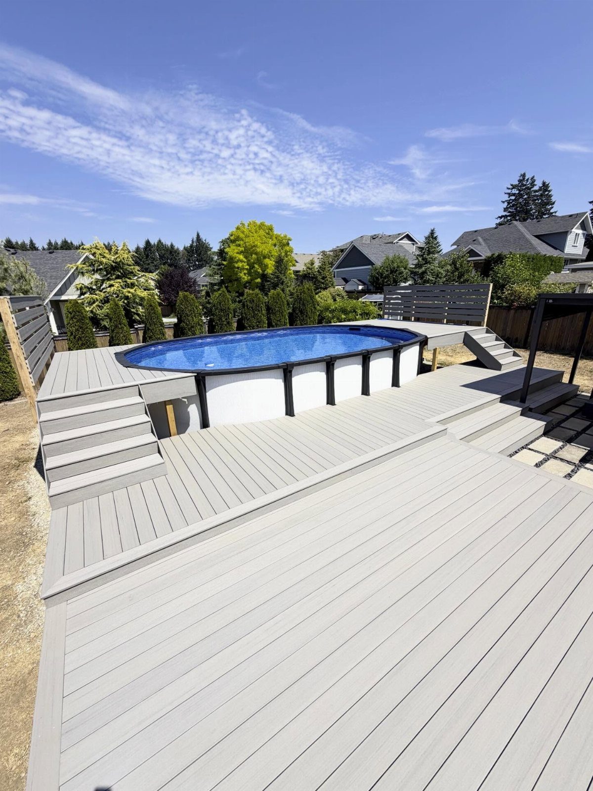 A light grey multi-level composite deck surrounding a blue above-ground pool in a backyard.