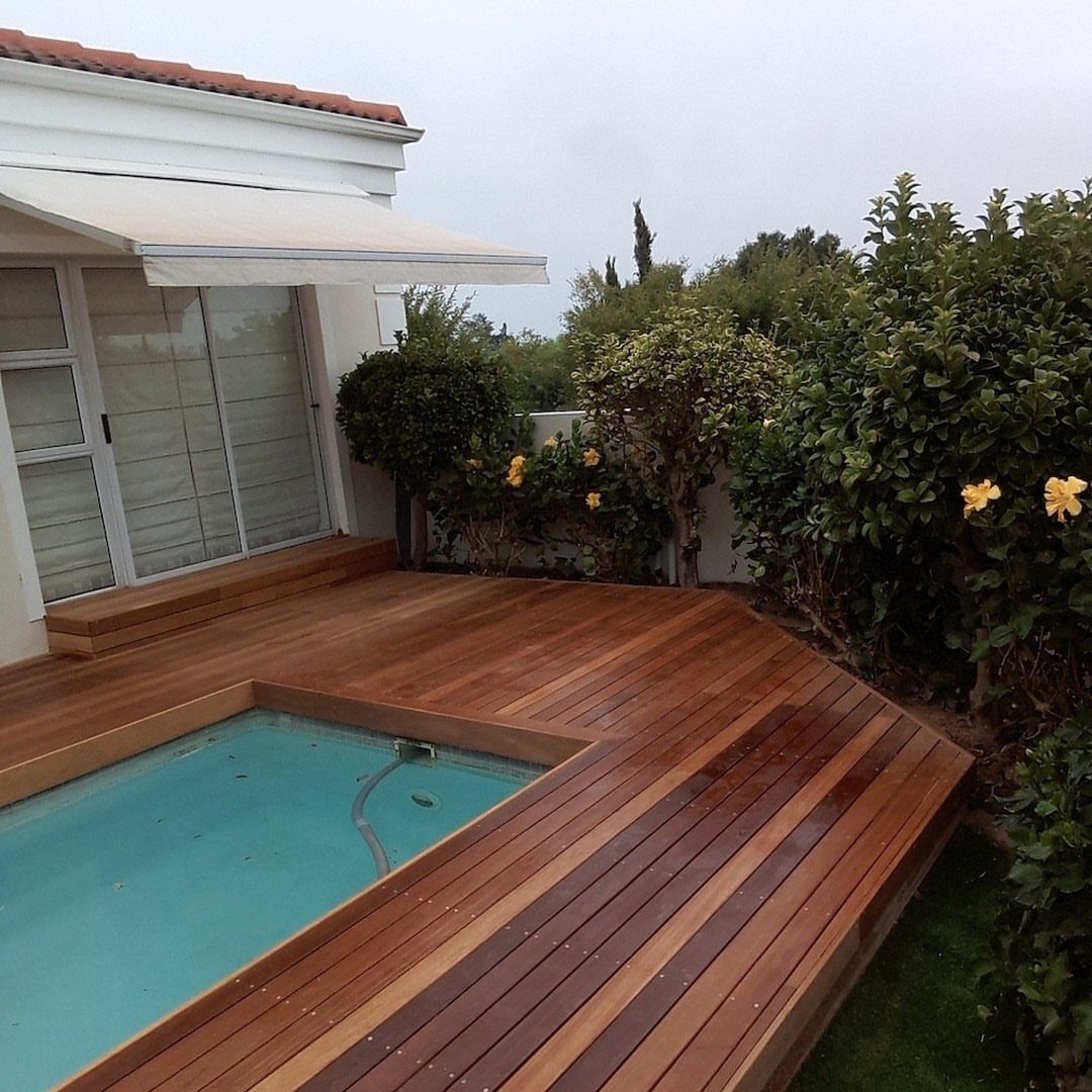 Polished wooden deck planks surrounding a bright blue swimming pool next to a white house.