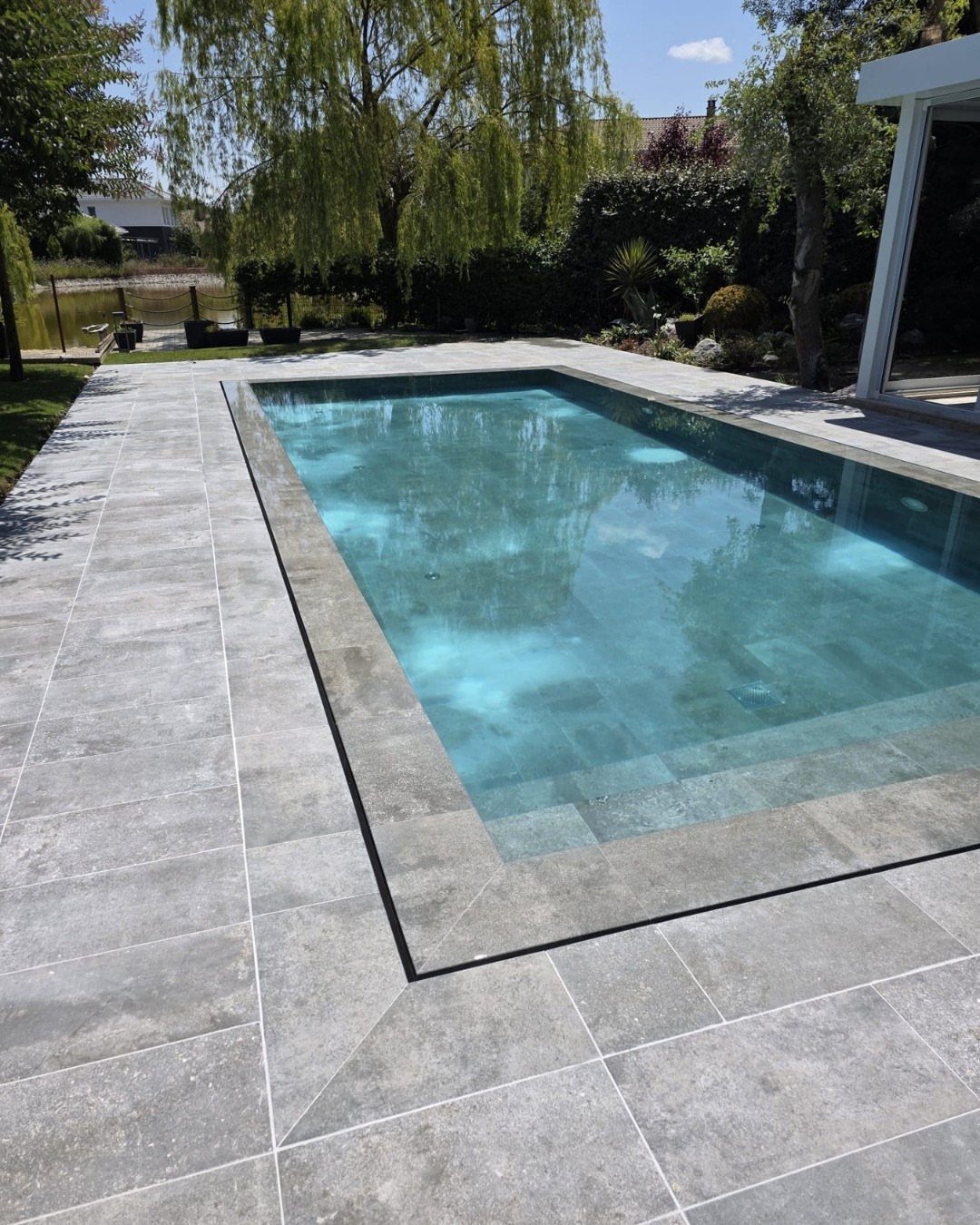A rectangular swimming pool surrounded by large grey stone tiles in a lush green backyard.