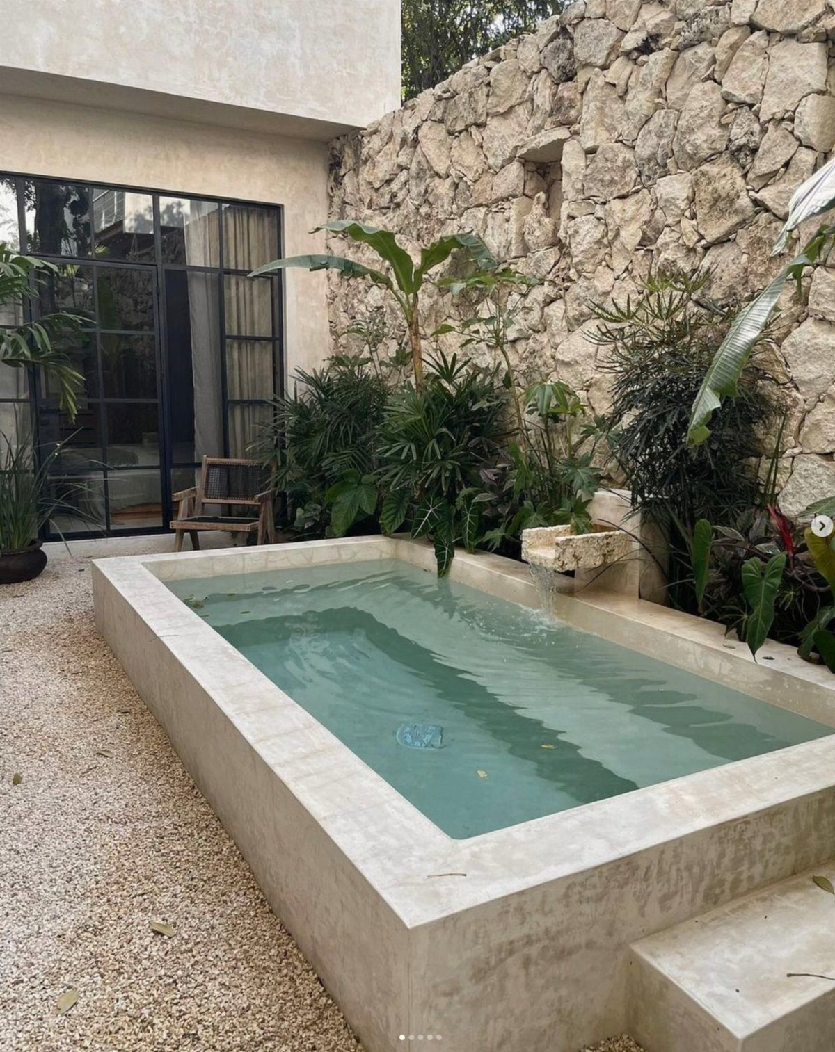 A rectangular concrete plunge pool surrounded by light pea gravel and lush tropical greenery against a stone wall.