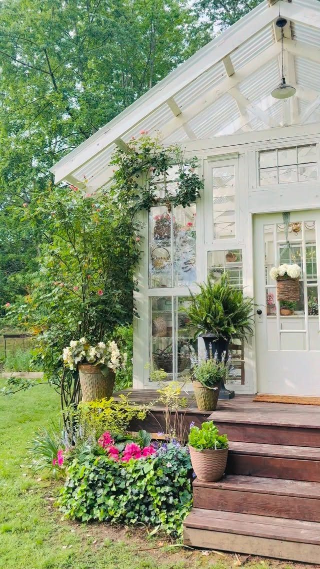 Cottage Garden Sheds with Porch: 13 Layouts and Looks 10 White wooden cottage shed with large windows, a wooden porch, and lush green garden plants.