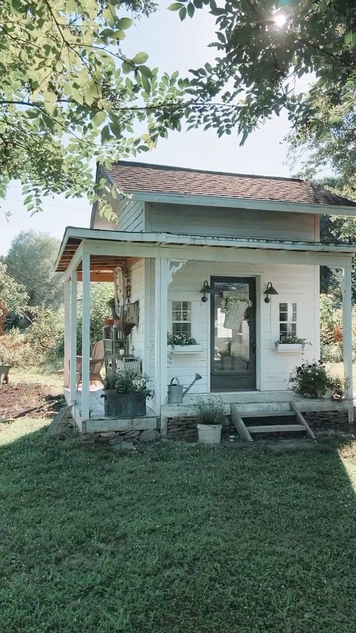 Cottage Garden Sheds with Porch: 13 Layouts and Looks 2 White wooden cottage shed with a covered porch, small windows, and steps in a garden.