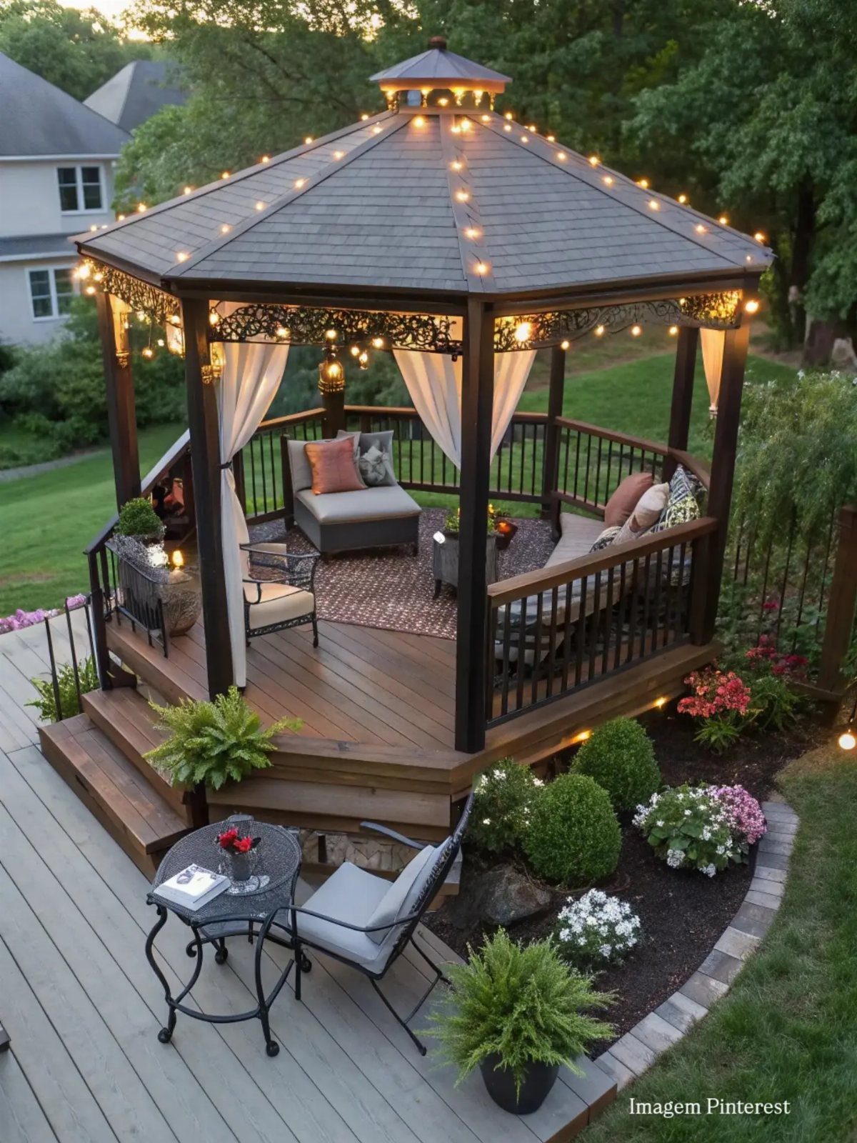 A dark wood gazebo with string lights and white curtains sits on a wooden deck.