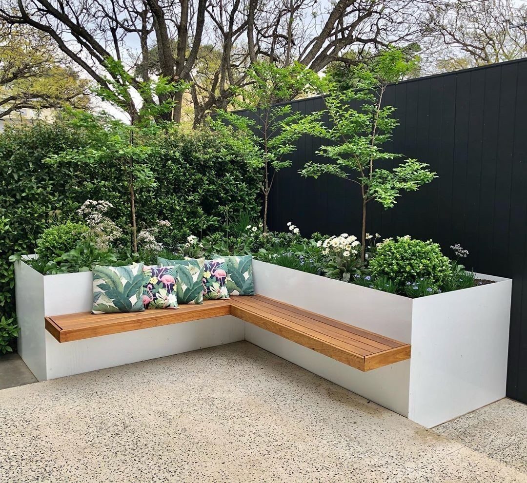 L-shaped white planter box with wooden bench seating and green plants on a stone patio.