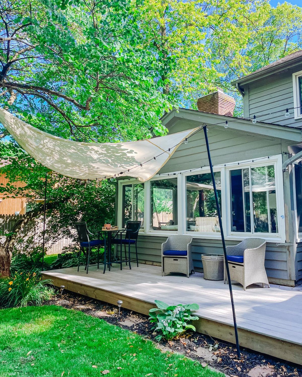 A cream triangular shade sail stretched over a wooden deck with outdoor furniture and green trees.