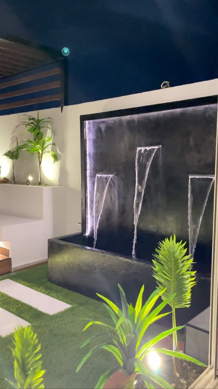 Black water feature wall with three cascading water streams into a dark basin surrounded by green plants.