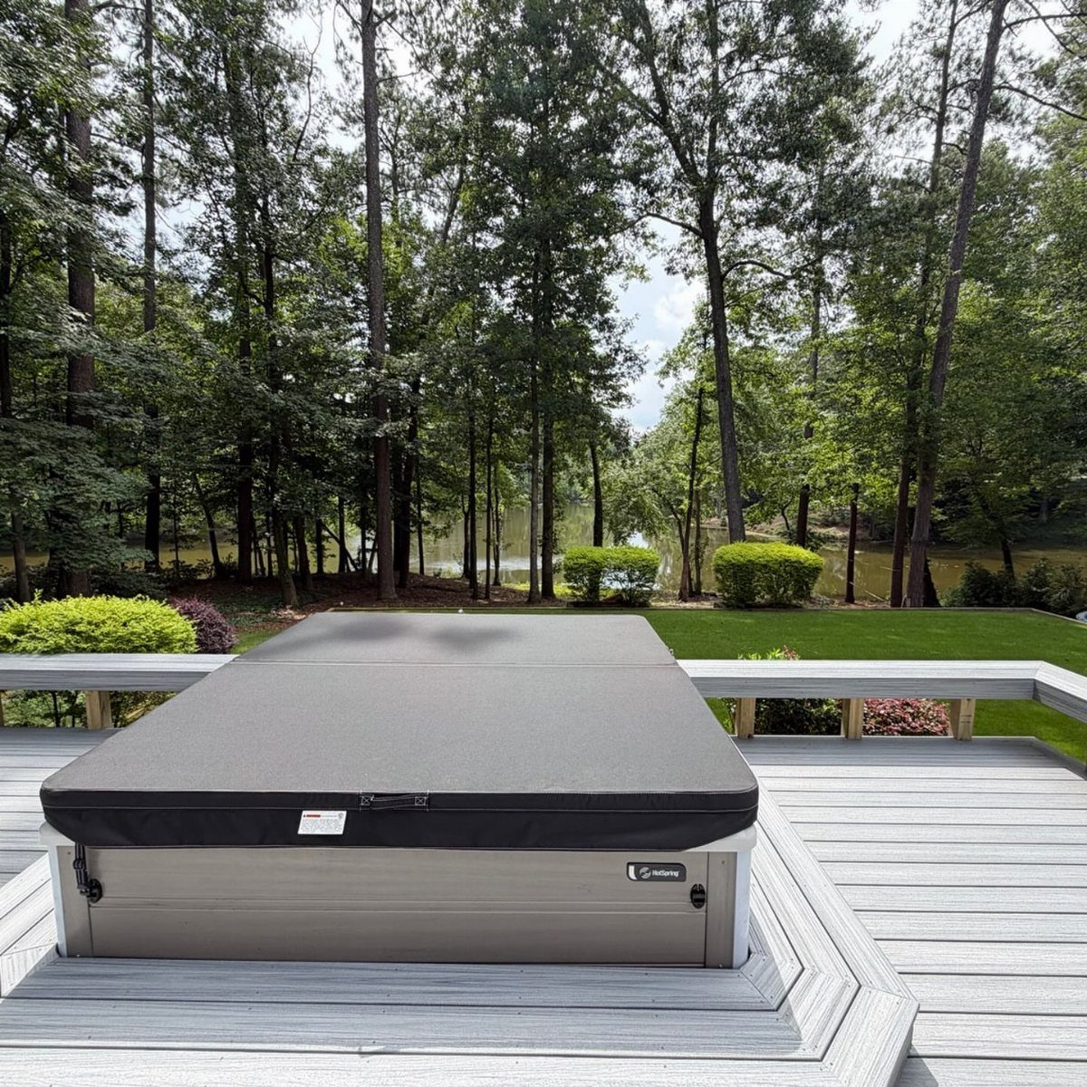 A grey composite deck featuring a large, covered hot tub overlooking a wooded lake area.