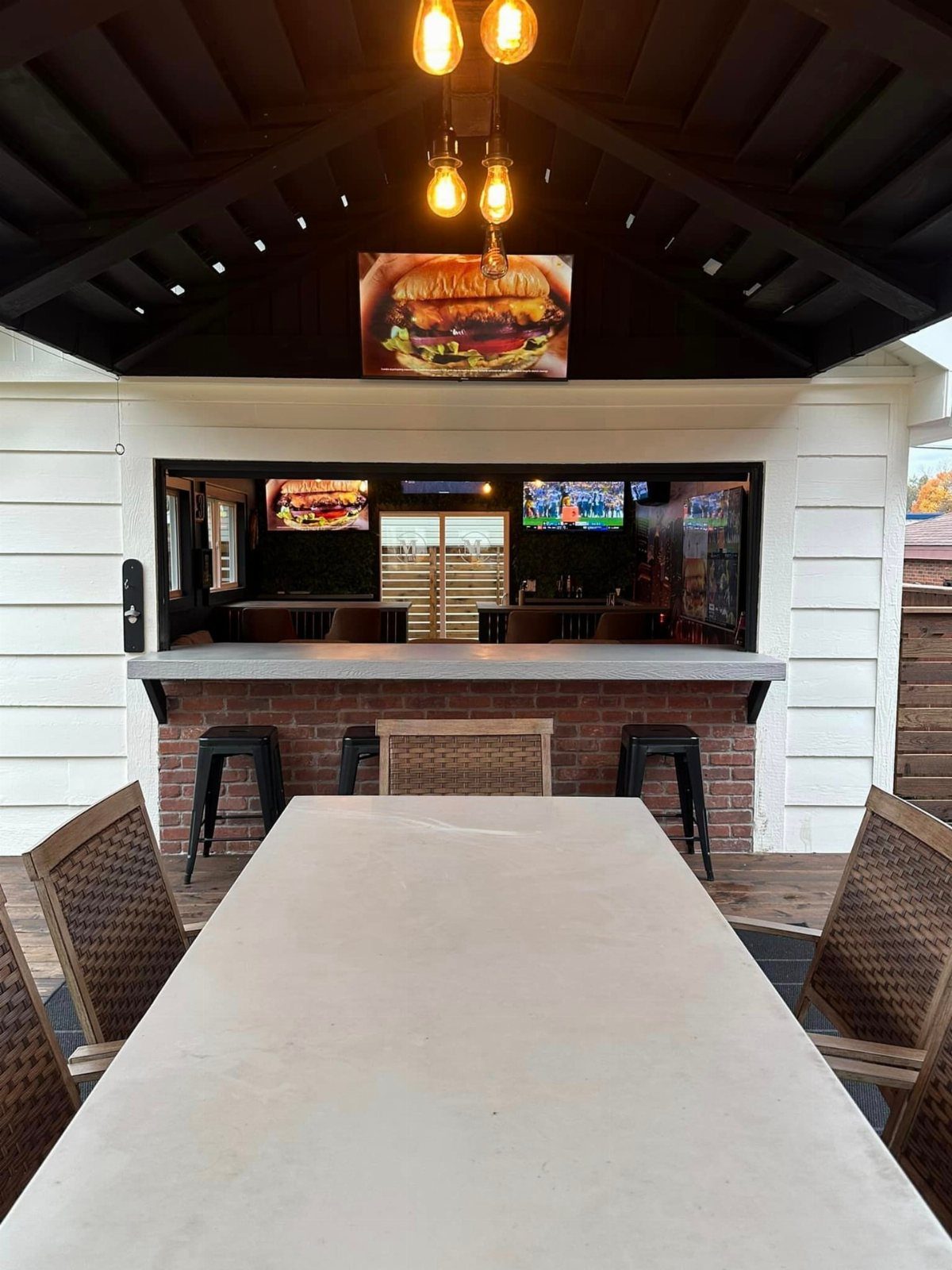 Outdoor bar with brick counter, black stools, and a long white dining table under a dark ceiling.