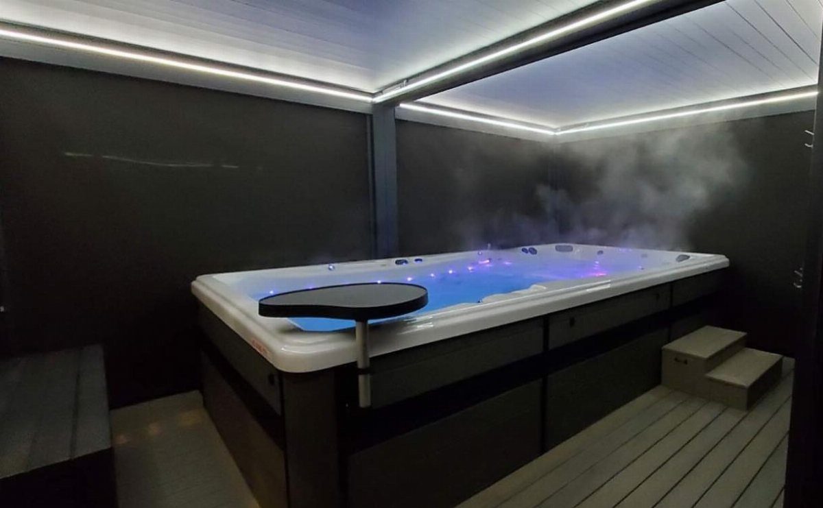 A steaming white hot tub with blue lights inside a dark, modern enclosed structure.