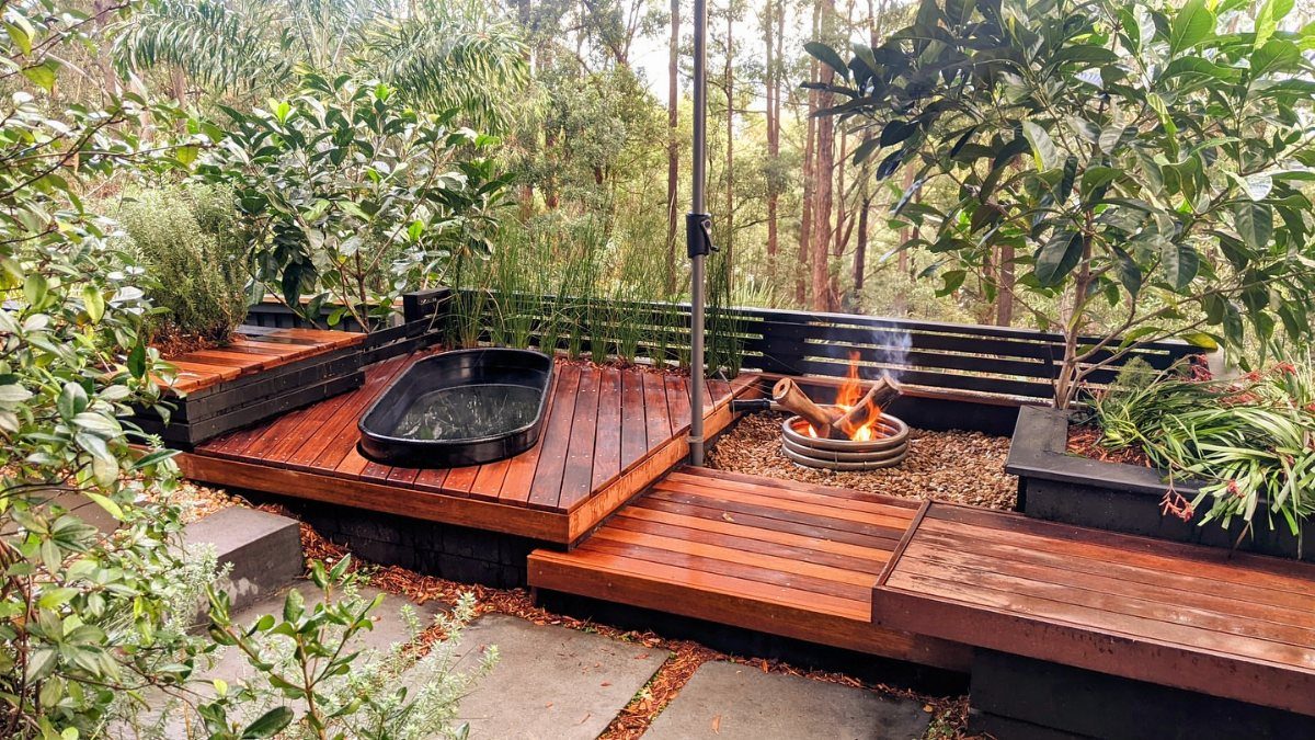 Wooden deck with a black soaking tub and fire pit nestled among green trees.