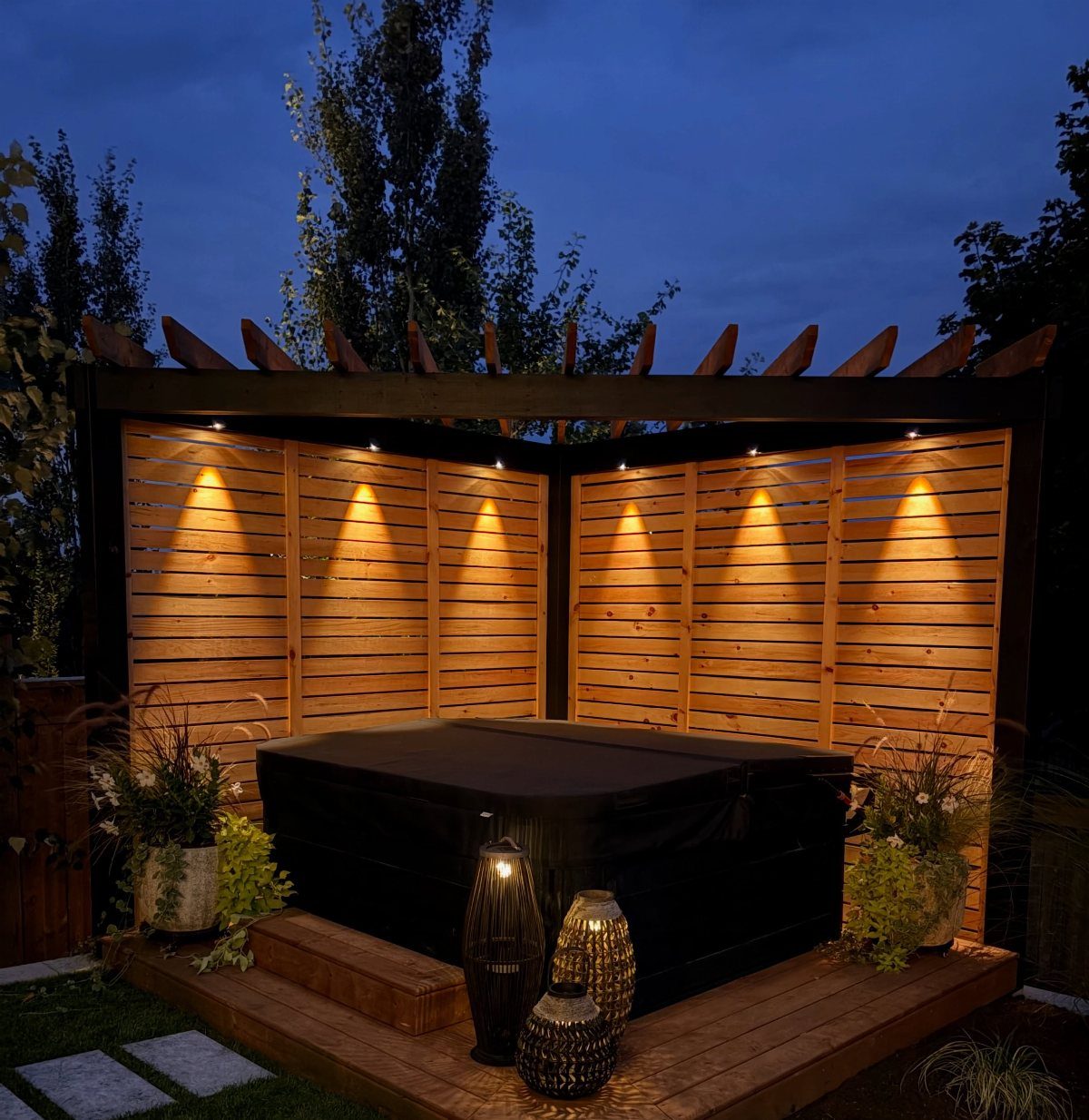 A black hot tub nestled within a wooden slat enclosure under a pergola at night.