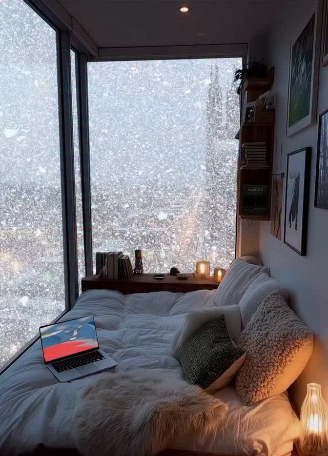 12 Decor for Winter After Christmas Ideas for a Cozy Home 8 A cozy bed with white linens and pillows sits next to a large window overlooking falling snow.