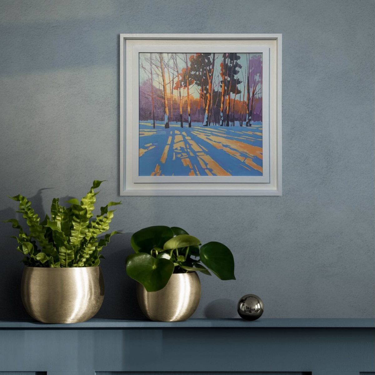 12 Decor for Winter After Christmas Ideas for a Cozy Home 10 Framed winter landscape painting above two gold potted plants on a blue console table.