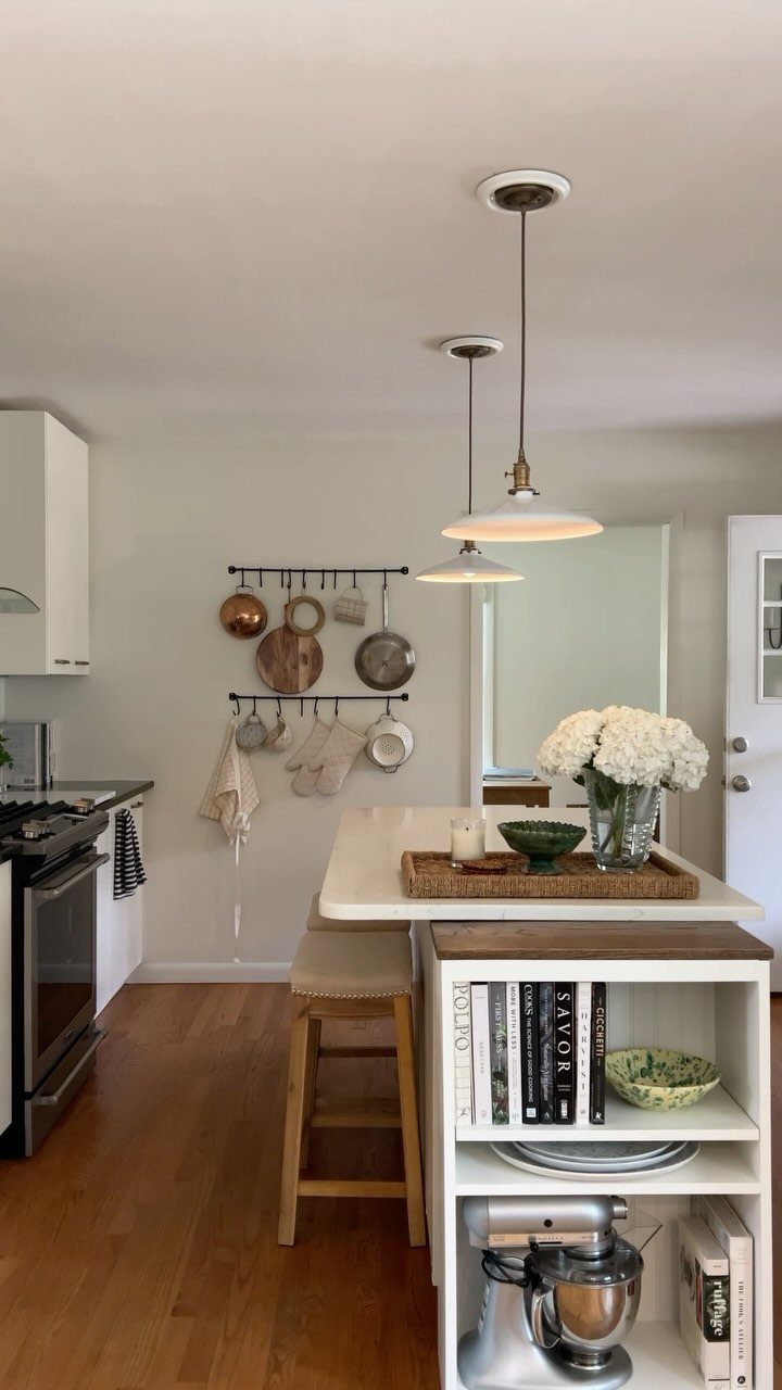 17 DIY Kitchen Island Ideas From Dressers to Live-Edge Slabs 4 White bookshelf kitchen island with cookbooks, flowers, and a stand mixer in a bright kitchen.