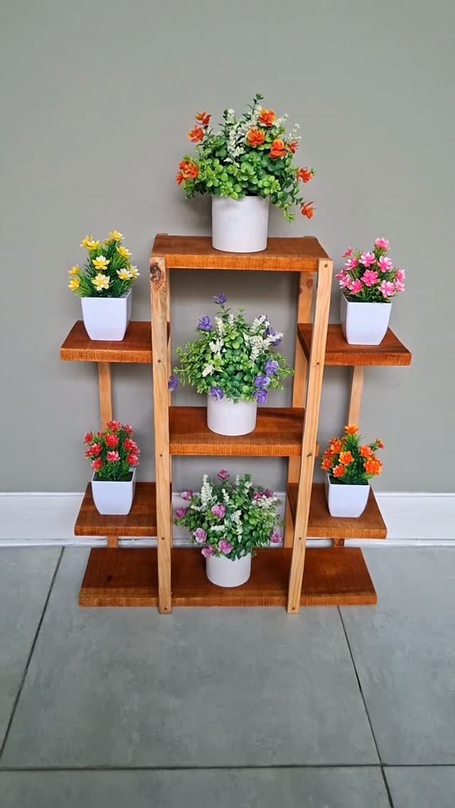 A multi-tiered wooden plant stand holding six white pots with colorful flowers against a grey wall.