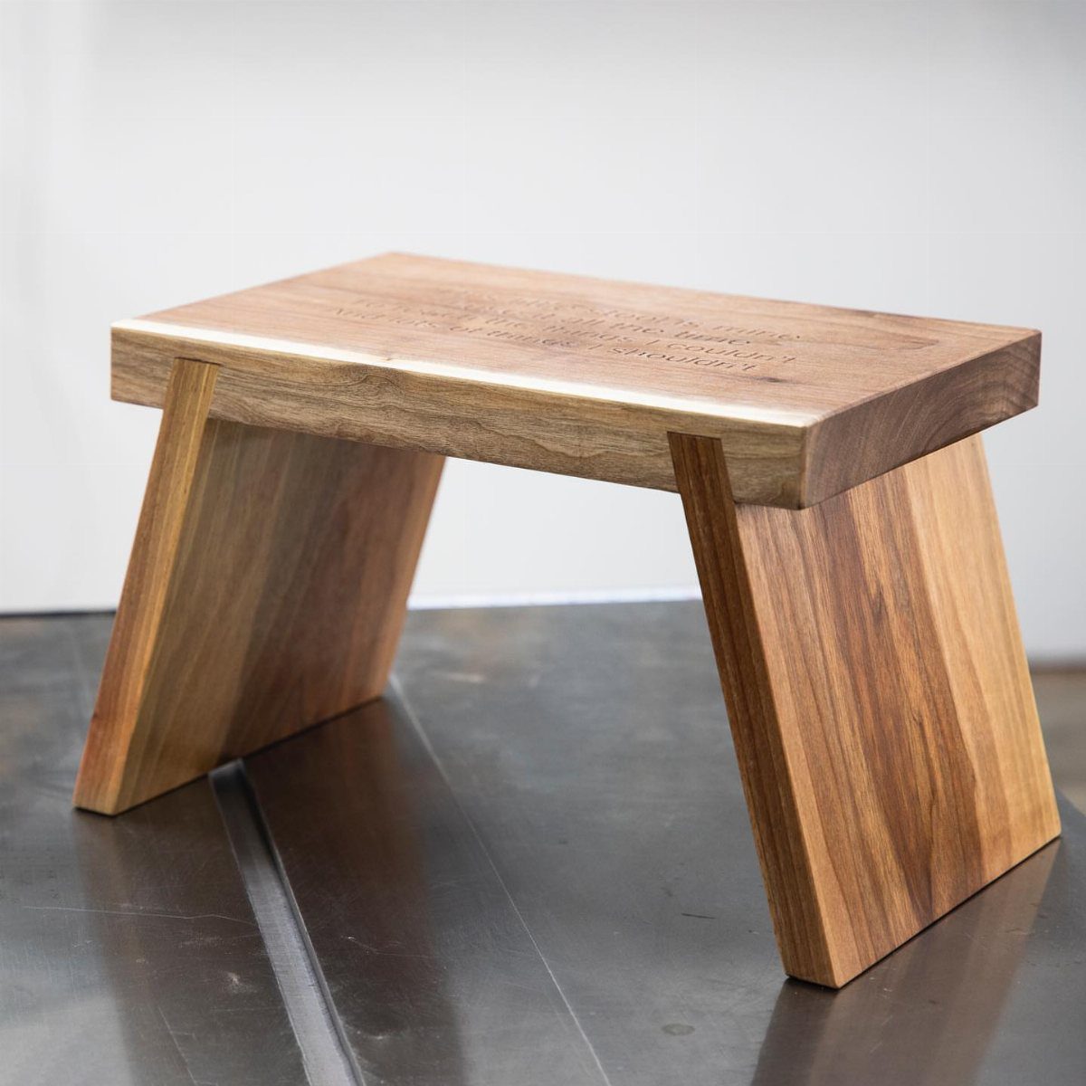 A minimalist wooden stool with thick planks and wide angled legs on a reflective floor.