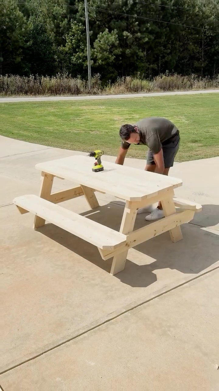 A man using a power drill to assemble a light wooden picnic table outdoors.