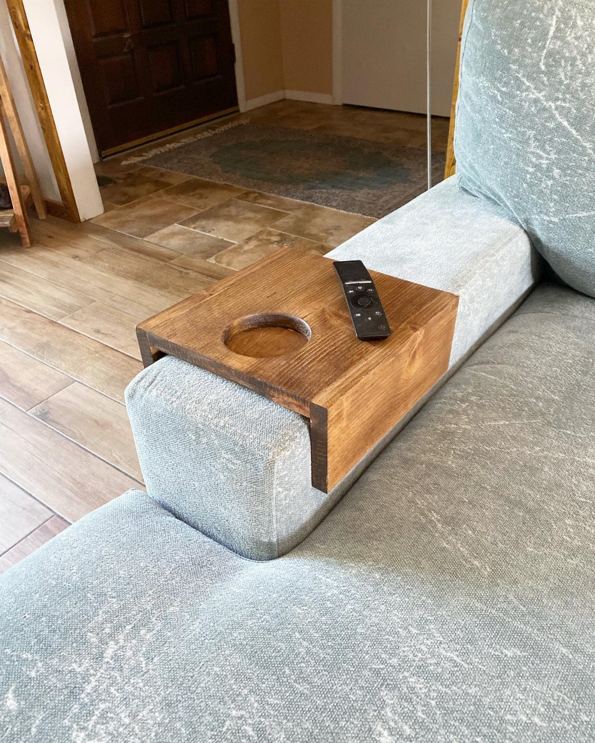 A dark stained wooden tray with a cup holder resting on a light blue sofa arm.