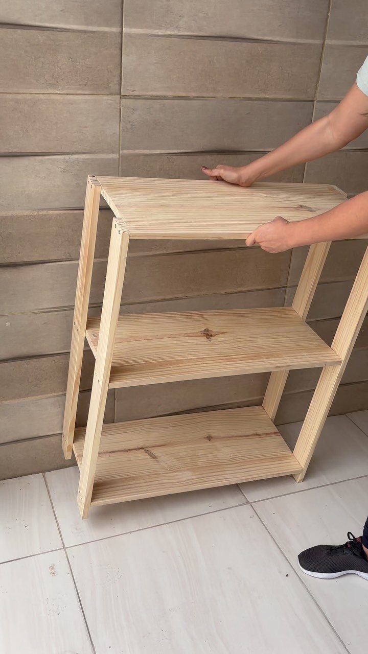 A person adjusts a three-tier light wood shelving unit against a textured wall.