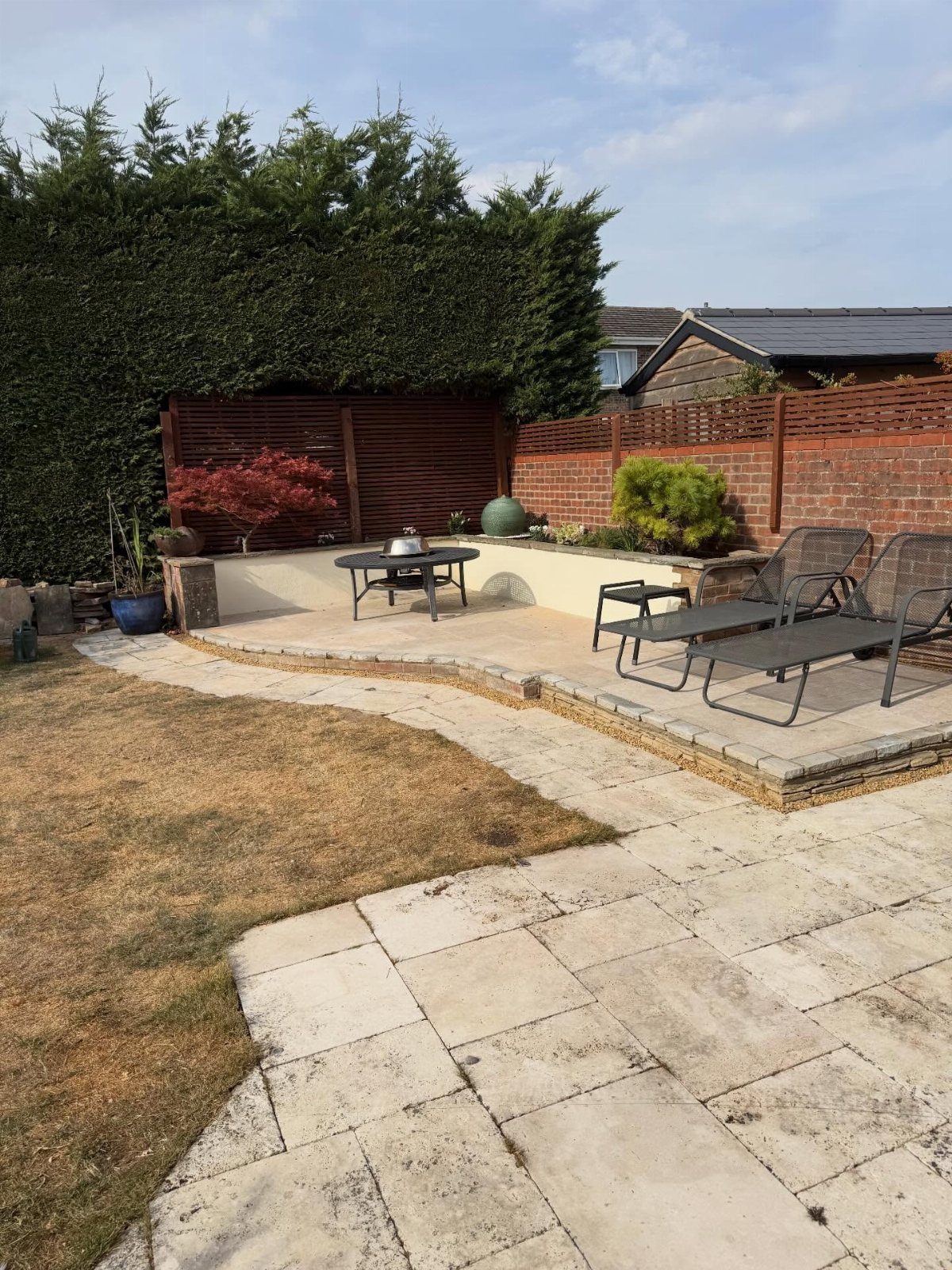 A stone patio with a raised seating area, lounge chairs, and a low retaining wall in a backyard.