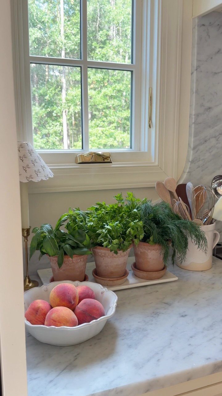 Three terracotta pots with green herbs sitting on a marble kitchen countertop near a window.