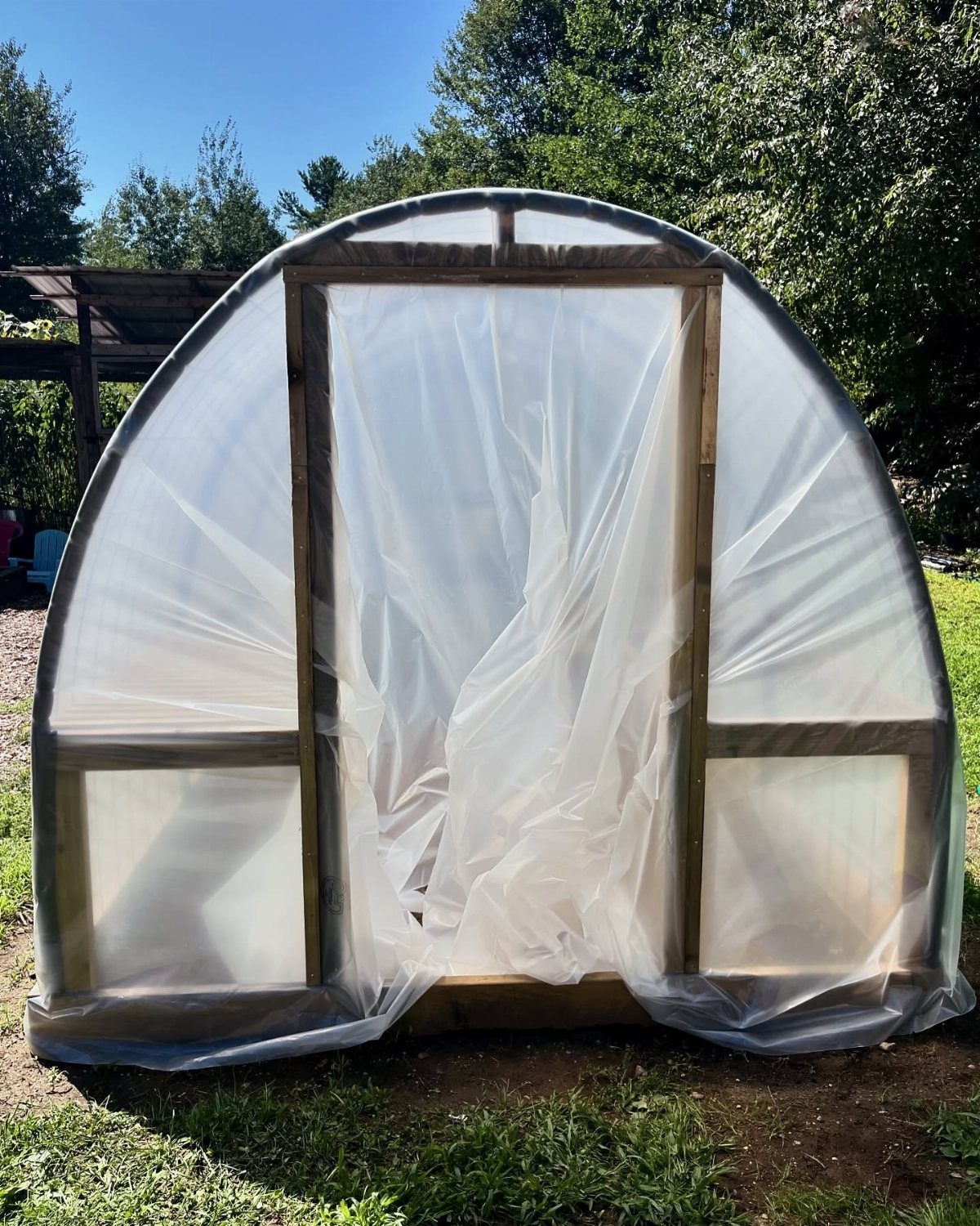 10 Easy Greenhouse Ideas for Any Backyard and Budget 9 A DIY arched greenhouse structure with a wooden frame and translucent plastic covering sitting on grass.