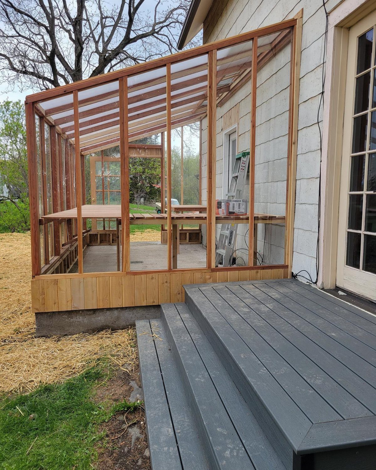10 Easy Greenhouse Ideas for Any Backyard and Budget 1 A wooden lean-to greenhouse frame under construction against a white house wall next to a deck.