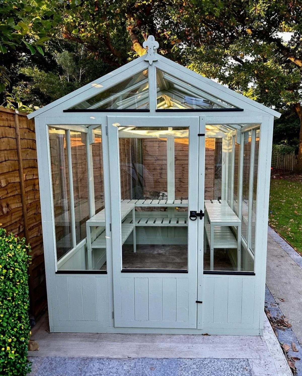 10 Easy Greenhouse Ideas for Any Backyard and Budget 7 A light sage green wooden greenhouse with glass panels sitting on a stone patio.