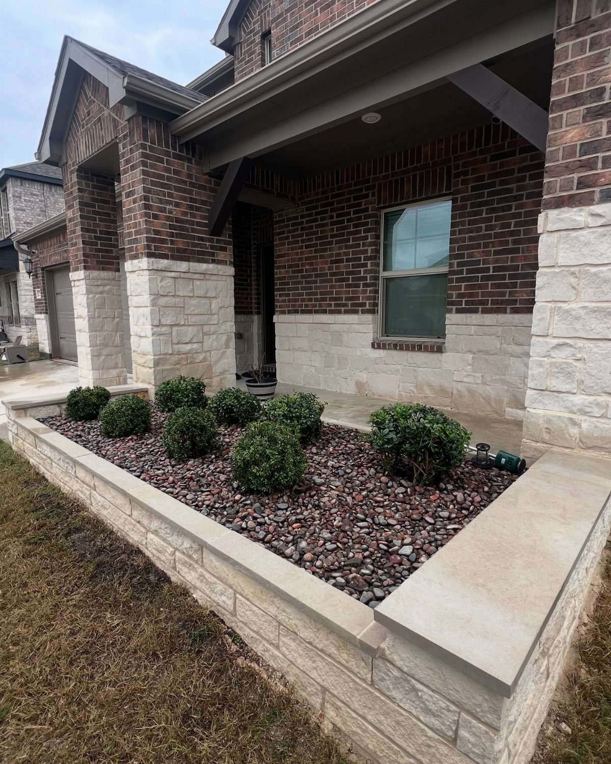 17 Front Flower Bed Ideas for Small Yards and Sunny Corners 11 Small green shrubs planted in a pebble-filled garden bed enclosed by a light stone masonry wall.