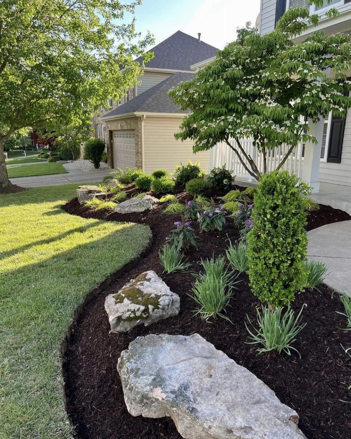 13 Front Lawn Landscaping Ideas for Curb Appeal and Low Upkeep 11 Landscaped garden bed with dark mulch, large rocks, green shrubs, and purple flowers near a house.