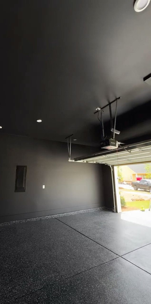 Black painted garage walls and ceiling with a dark speckled epoxy floor and open garage door.