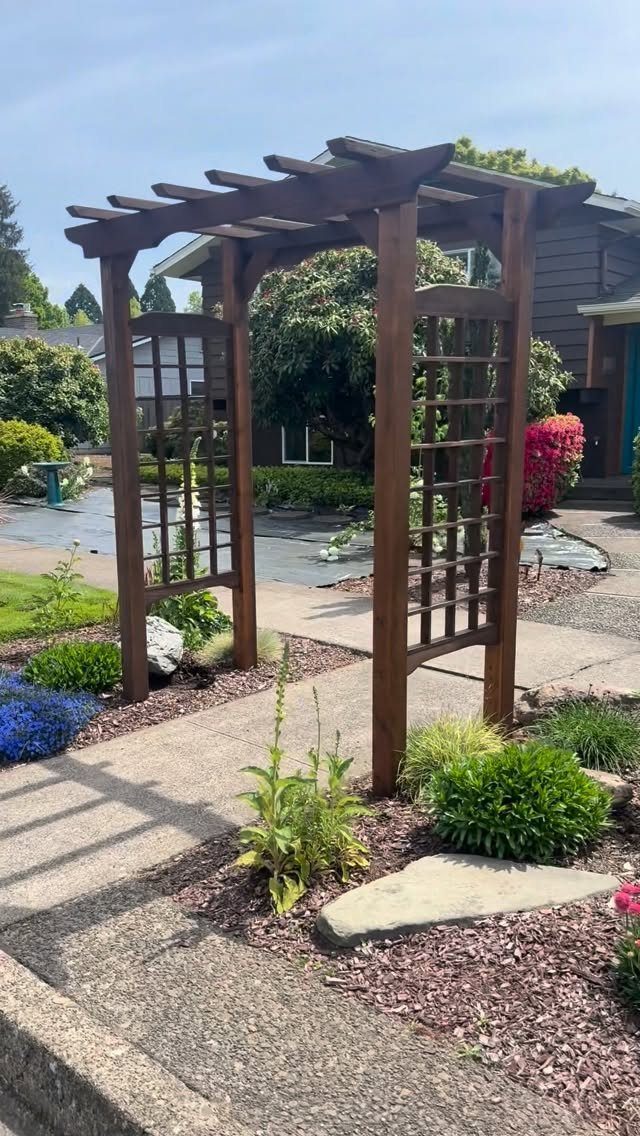 10 Garden Front of House Ideas: From Foundation Beds to Arbors 7 A dark wooden arbor with lattice sides stands over a concrete walkway in a garden.