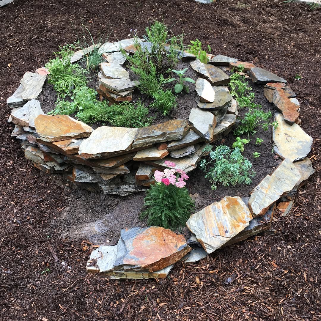 A circular herb spiral constructed from layered flat stones with green plants growing inside.