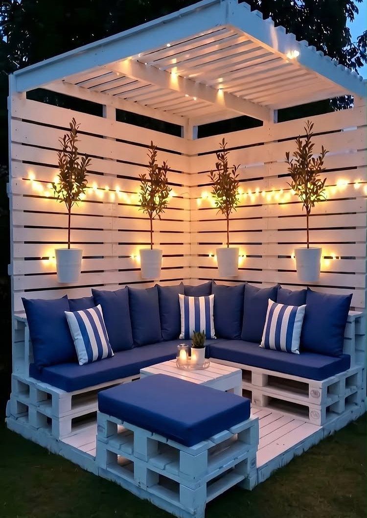 16 Low Budget Backyard Ideas to Refresh Your Outdoor Space 14 White pallet furniture with blue cushions and string lights under a wooden pergola outdoors.