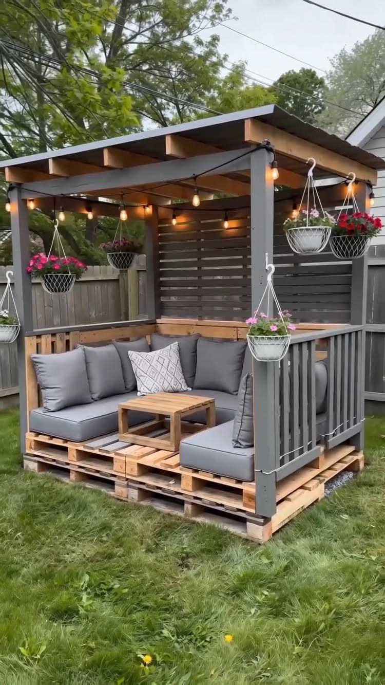 16 Low Budget Backyard Ideas to Refresh Your Outdoor Space 6 A backyard lounge area constructed from wooden pallets with grey cushions and string lights.
