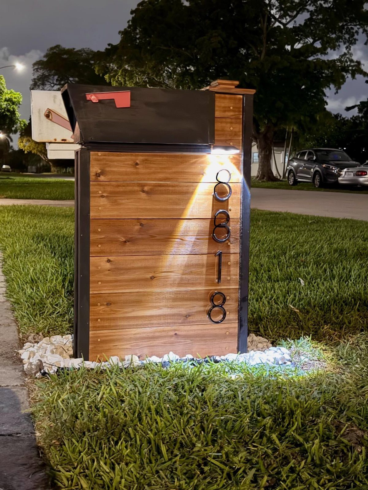 Modern wooden mailbox stand with black metal accents and house numbers on a grassy lawn.