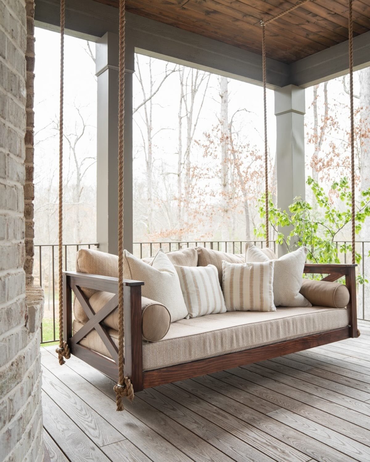 A wooden porch swing with beige cushions and pillows hangs on a covered porch.
