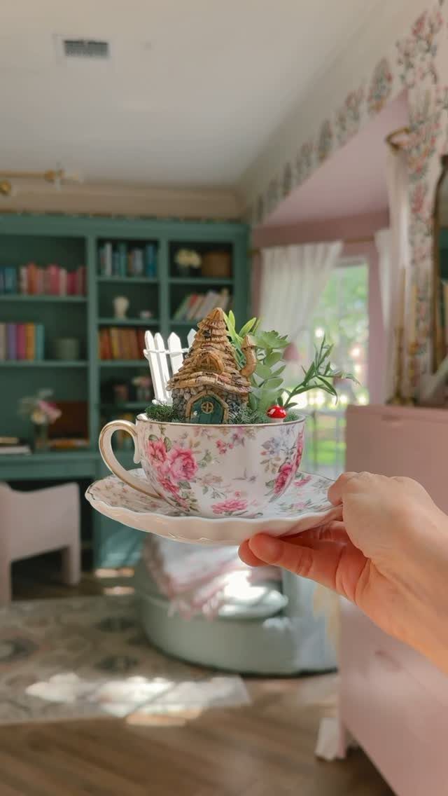 A hand holds a floral teacup containing a miniature fairy garden with a tiny house.