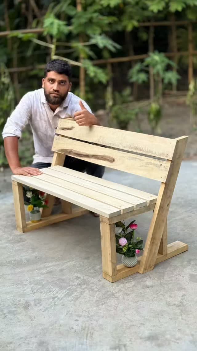 A man sitting behind a handmade wooden bench with small potted flowers at its base.