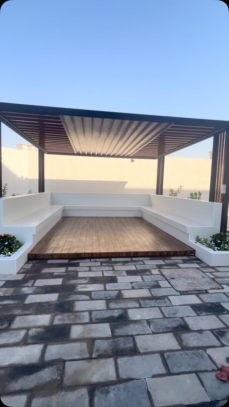 18 Yard Layout Ideas with Photos for Front and Back Spaces 12 White built-in concrete benches surrounding a wooden deck under a dark pergola structure.