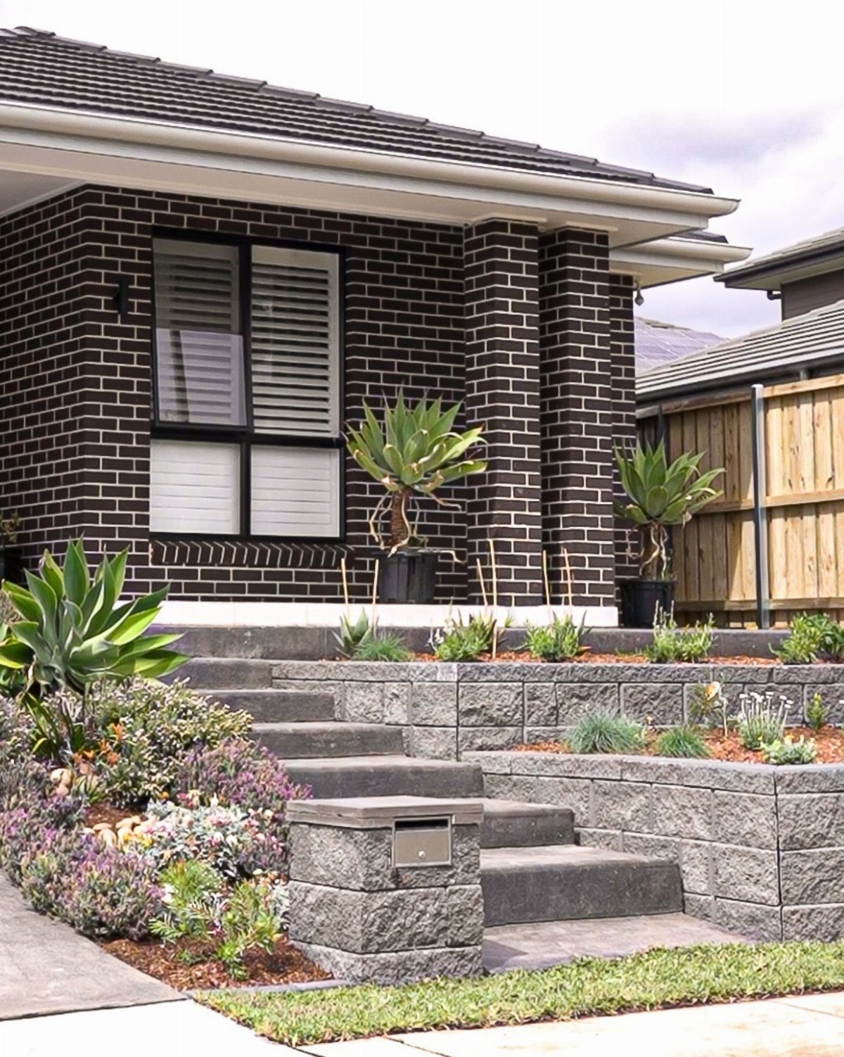 18 Yard Layout Ideas with Photos for Front and Back Spaces 15 Grey stone retaining walls create tiered garden levels with succulents in front of a dark brick house.